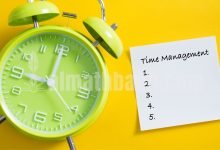 Jam alarm berwarna hijau dengan kertas catatan kecil bertuliskan "management", ilustrasi pengingat manajemen waktu.